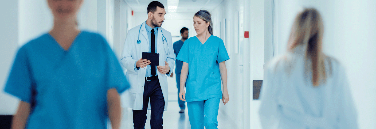 Ein Arzt im weißen Kittel spricht mit einer Krankenschwester in blauer Kleidung, während sie gemeinsam einen Krankenhausflur entlanggehen. Beide wirken konzentriert und scheinen ein wichtiges Thema zu besprechen.